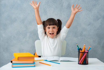 Back to school. Cute child schoolgirl sitting at a Desk in the room. The kid is learning and doing his homework. The girl is emotionally happy with her hands up.